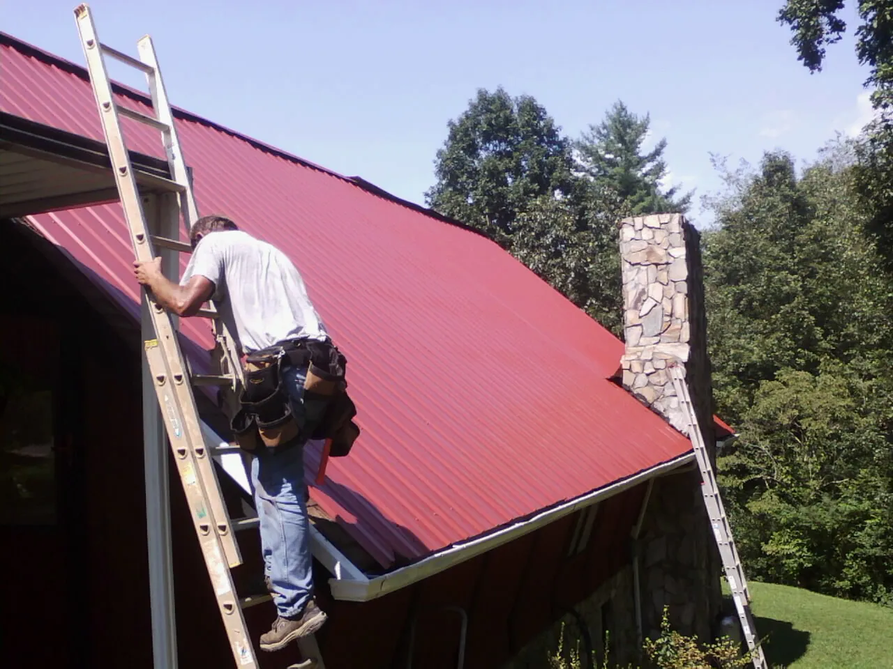 Roofer on a ladder beside a red metal roof and stone chimney — Soffit & Fascia Repair by Roofing Brothers in Green Bay